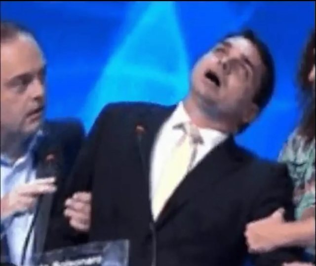 Flávio Bolsonaro passando mal no debate da Band em 2016, de olhos fechados e boca aberta, amparado por outros candidatos